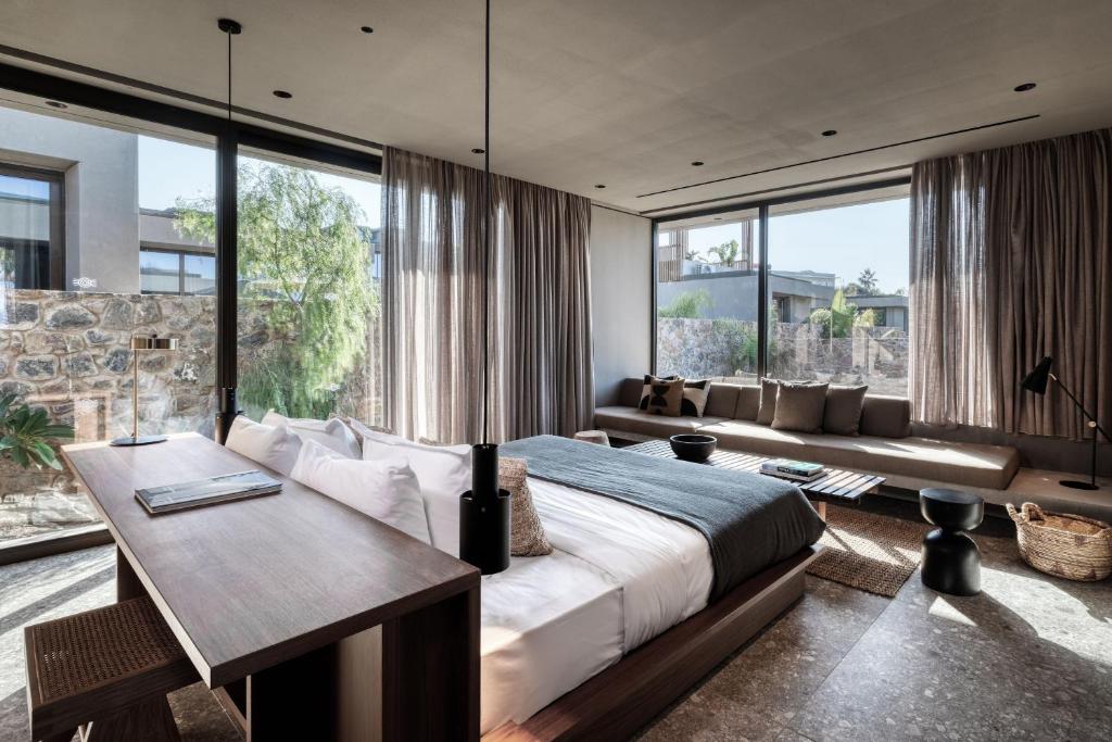 Mūsdienīga guļamistaba ar plašu logu, kas piedāvā skatu uz dārzu, ērtiem dīvāniem un stilīgu darba vietu. Interjers ir minimālistisks, ar dabiskām krāsām un tekstūrām.