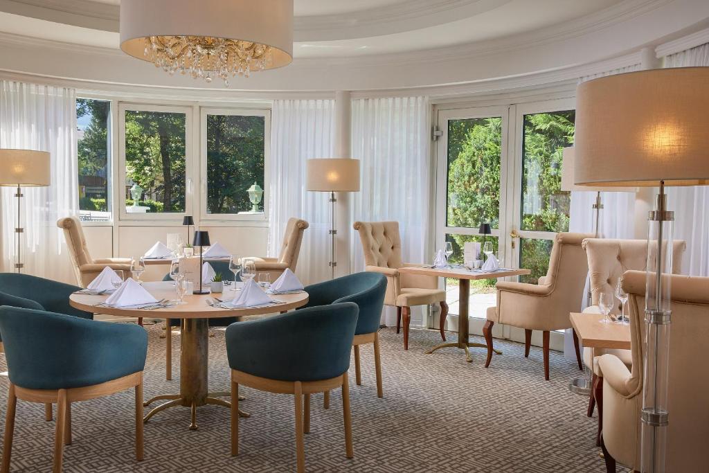 Elegants restorāna interjers ar apaļu galdu, zilu krēslu komplektu un gaismas spuldzēm, kas rada siltu atmosfēru. Lielie logi nodrošina dabisko apgaismojumu un skatu uz dārzu.