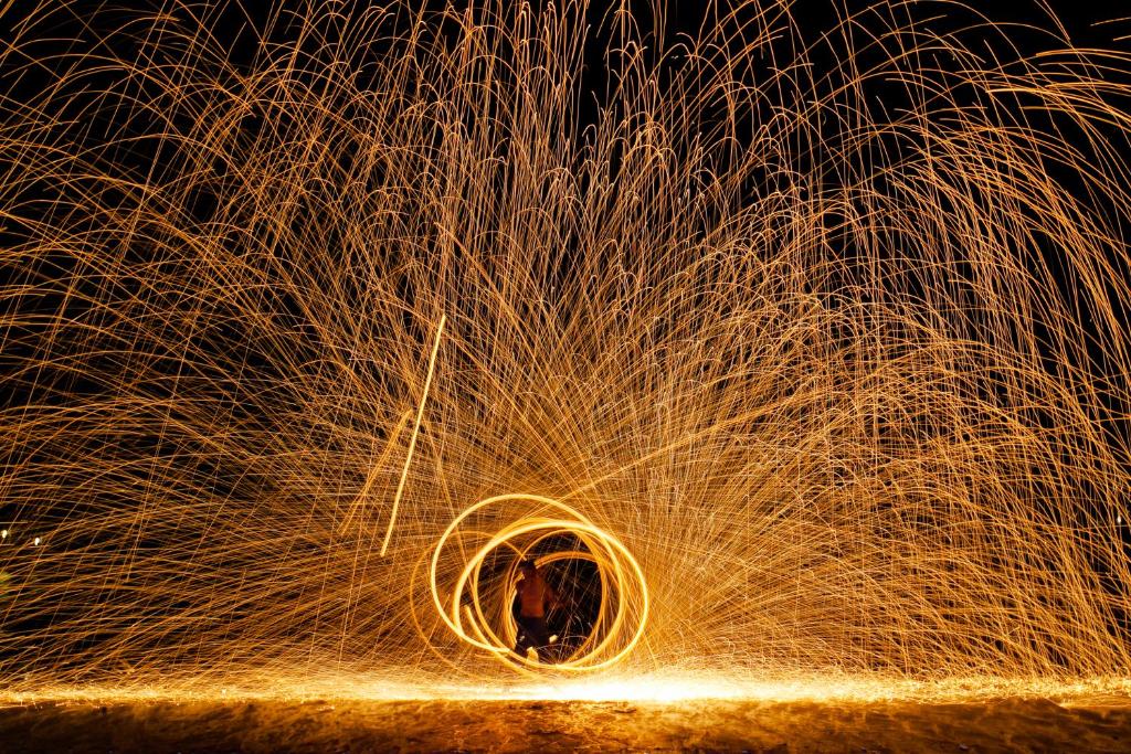 Uguns šova izpildītājs veido krāšņu dzirksteļu apli tumsā, radot dinamisku un aizraujošu skatu. Sarkanās un dzeltenās dzirksteles izplatās visapkārt, radot maģisku atmosfēru.