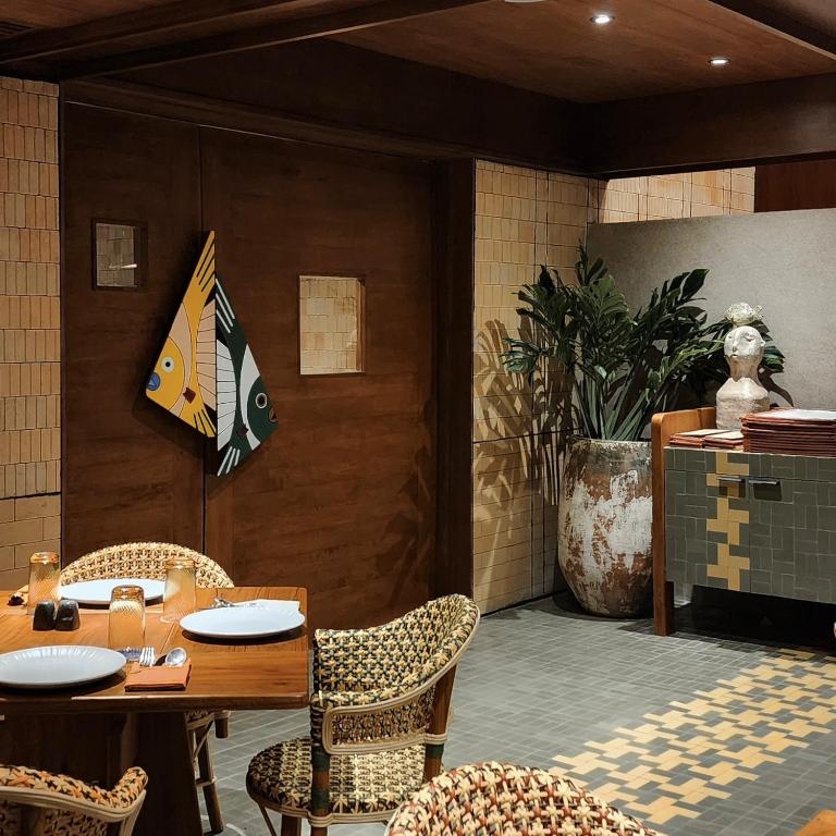 Mūsdienīgs restorāna interjers ar koka durvīm, dekorēts ar krāsainu karogu, augiem un mākslas elementiem, kas rada siltu un viesmīlīgu atmosfēru.