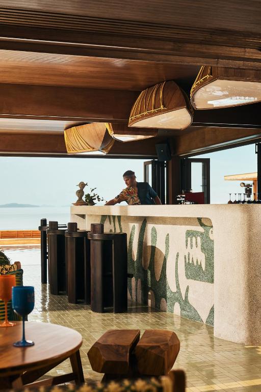 Mūsdienīgs bārs ar koka detaļām un māksliniecisku izskatu, kurā redzams bārmenis, kas pasniedz dzērienus. Fons ietver mierīgu jūras ainavu un stilīgas mēbeles.