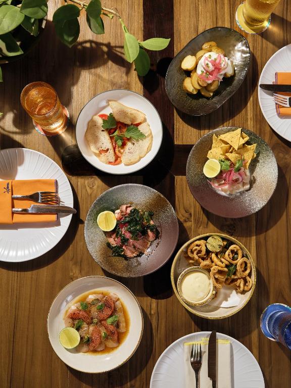 Dažādu ēdienu izlase, kas pasniegta uz koka galda, ietver tacos, čipsus ar guakamoli, ceptus kalmārus un salātus. Galda dekors papildina dzērieni un augu elements.