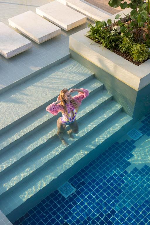 Sieviete ar rozā blūzi un peldkostīmu stāv ūdenī pie baseina kāpnēm, baudot sauli un atpūtu. Baseins ir moderns ar zilu flīzēm un augiem apkārt.