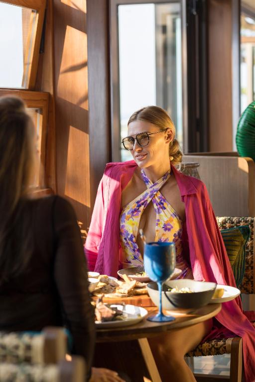 Sieviete ar saulesbrillēm un krāsainu apģērbu sēž pie galda, baudot maltīti un sarunājoties ar otru personu. Gaismas plūsma caur logiem rada siltu atmosfēru.