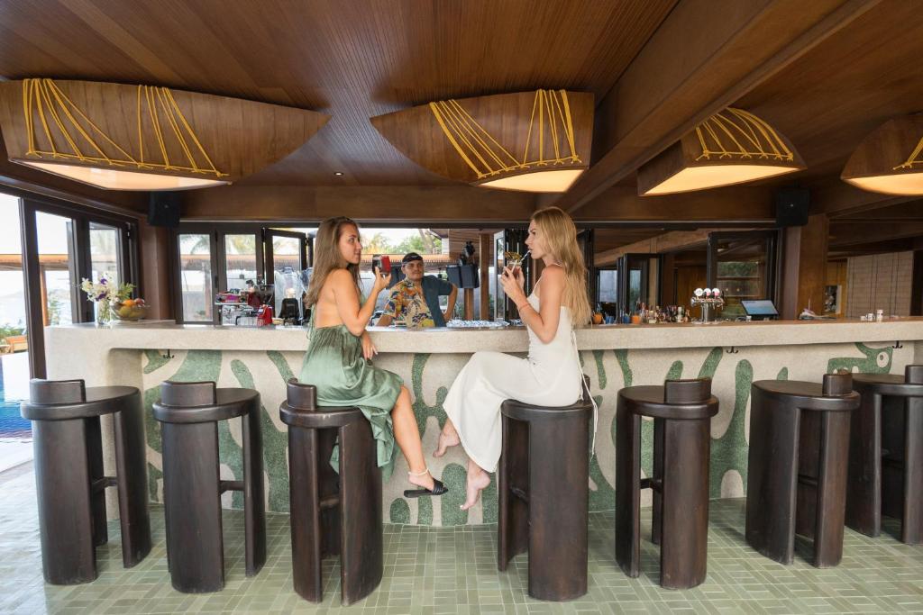 Divas sievietes sēž bārā, baudot dzērienus un sarunājoties, kamēr bārmenis stāv aiz letes. Interjērs ir moderns ar koka detaļām un stilīgām lampām, radot relaksējošu atmosfēru.