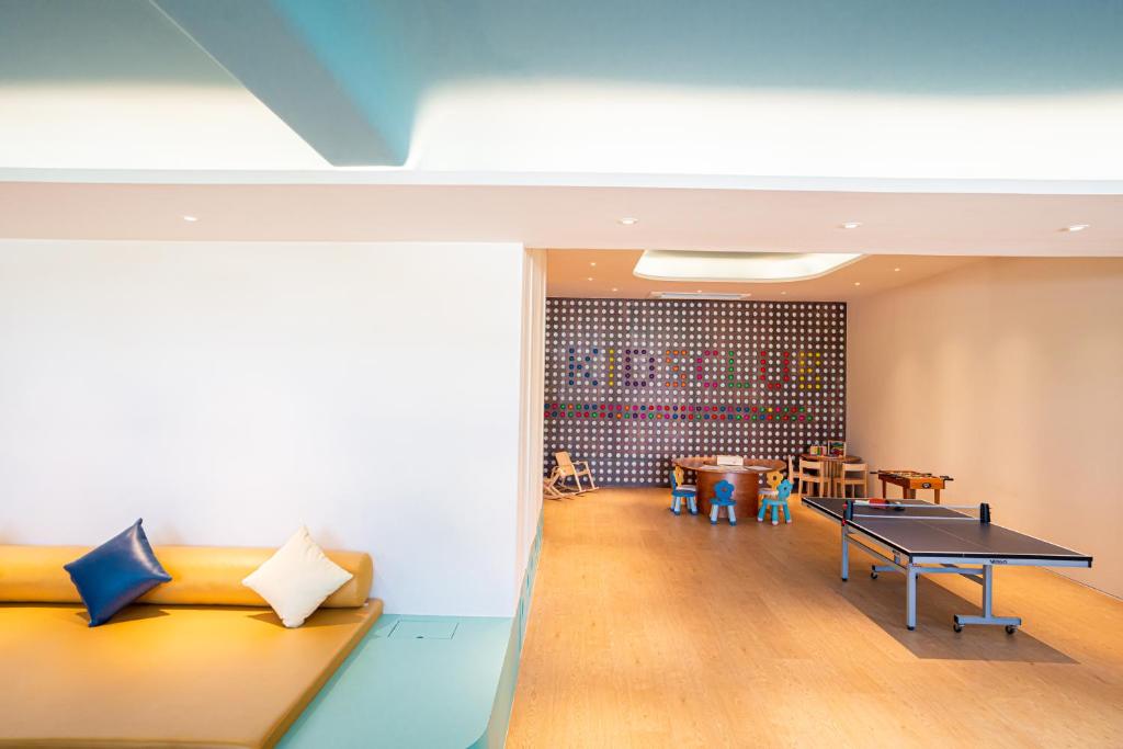 Mūsdienīga bērnu spēļu telpa ar pingponga galdu, krāsainiem krēsliem un interaktīvām aktivitātēm. Telpai ir gaiša un atvērta atmosfēra, kas veicina radošumu un spēlēšanu.