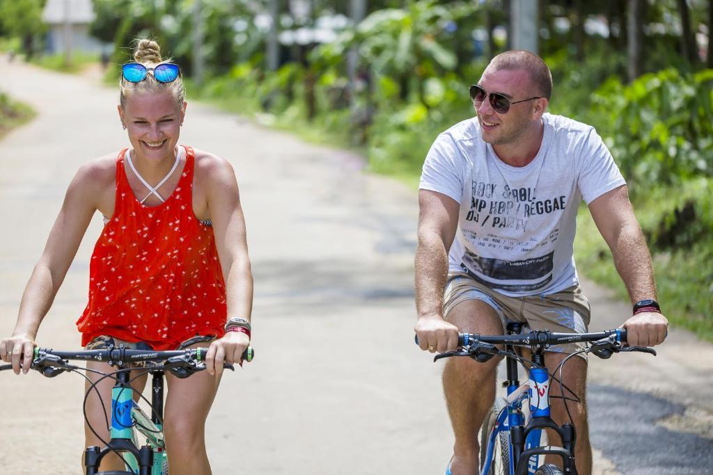 Divi cilvēki, vīrietis un sieviete, brauc ar velosipēdiem pa saulainu ceļu, baudot dabu un brīvdienu noskaņu. Viņi izskatās priecīgi un relaksēti.