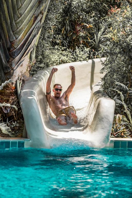 Vīrietis, kas ar paceltām rokām slīd pa ūdens slaidu, izsaka prieku un aizrautību, kamēr viņš ieiet baseinā. Apkārt ir tropiska vide ar zaļām augiem un krāšņām ainavām.