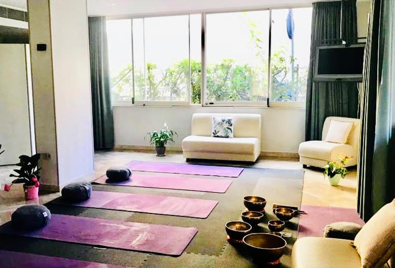 Gaiša un mierīga jogas zāle ar rozā paklājiem, akmeņiem un dziedinošām traukām, kas izkārtotas uz grīdas. Logi nodrošina dabīgu apgaismojumu un skatu uz zaļumu.