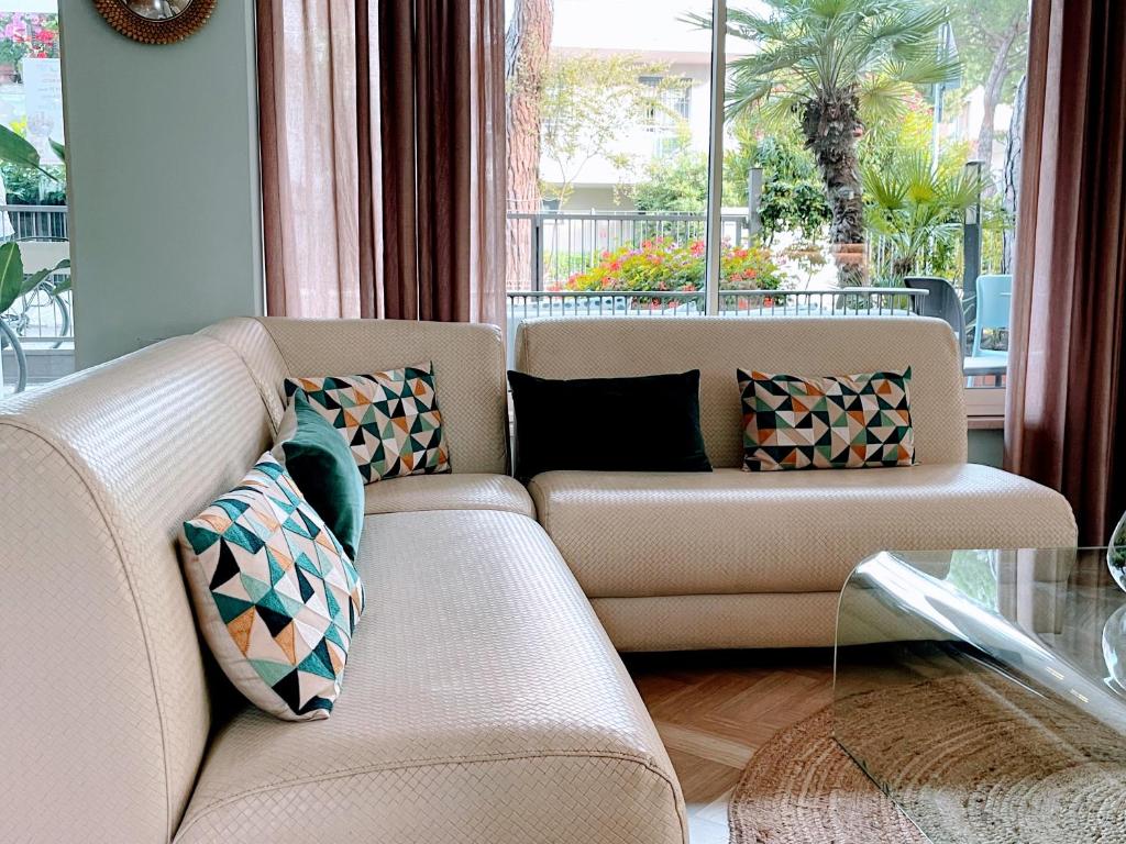 Mūsdienīgs viesistabas stūris ar gaišu dīvānu un ģeometriskām spilveniem, kas rada mājīgu atmosfēru. Logi atver skatu uz zaļu dārzu un palmu kokiem.