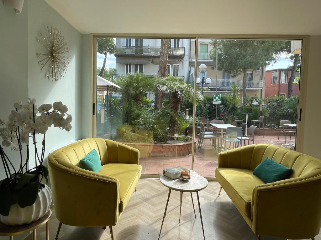 Gaiša un moderna telpa ar dzeltenām mēbelēm, kas vērsta uz dārzu ar palmām un ārējām sēdvietām. Logā redzama zaļā zāle un pilsētas ainava.