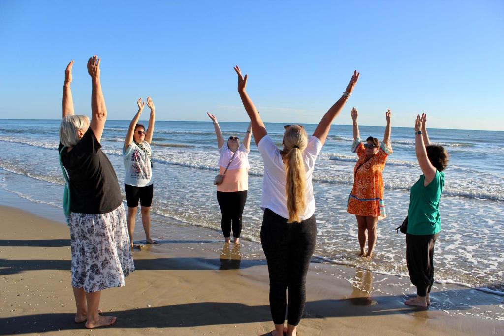 Grupas sieviešu, kas stāv uz pludmales smiltīm un veic vingrošanas kustības ar rokām gaisā, priecīgā atmosfērā pie jūras.