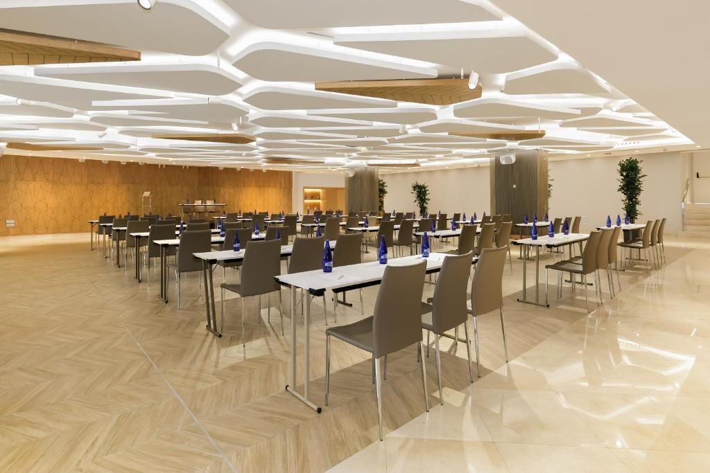 Mūsdienīgs konferenču zāles interjers ar rindu galdiem un krēsliem, apgaismojums ar ģeometriskām formām un dekoratīvām augiem. Ideāli piemērots uzņēmumu pasākumiem un sanāksmēm.