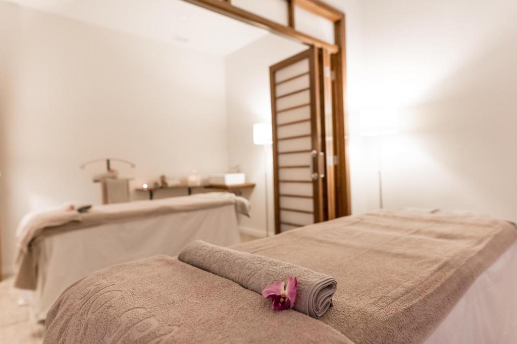 Mierīgā spa telpa ar masāžas galdiem, uz kuriem novietoti dvieļi un ziedi, radot relaksējošu atmosfēru. Interjerā ir mīkstas gaismas un eleganta dizaina elementi.