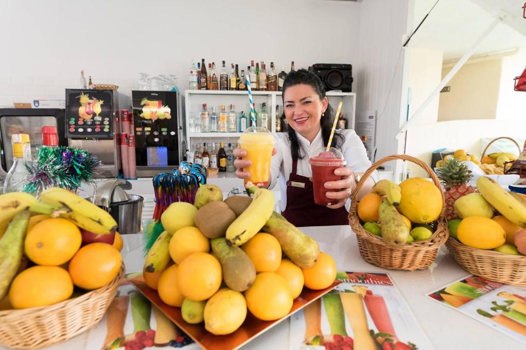 Svaigu augļu stends ar dzērieniem, kurā sieviete pasniedz divus kokteiļus. Fons rotāts ar alkoholiskajiem dzērieniem un augļiem, radot patīkamu atmosfēru.