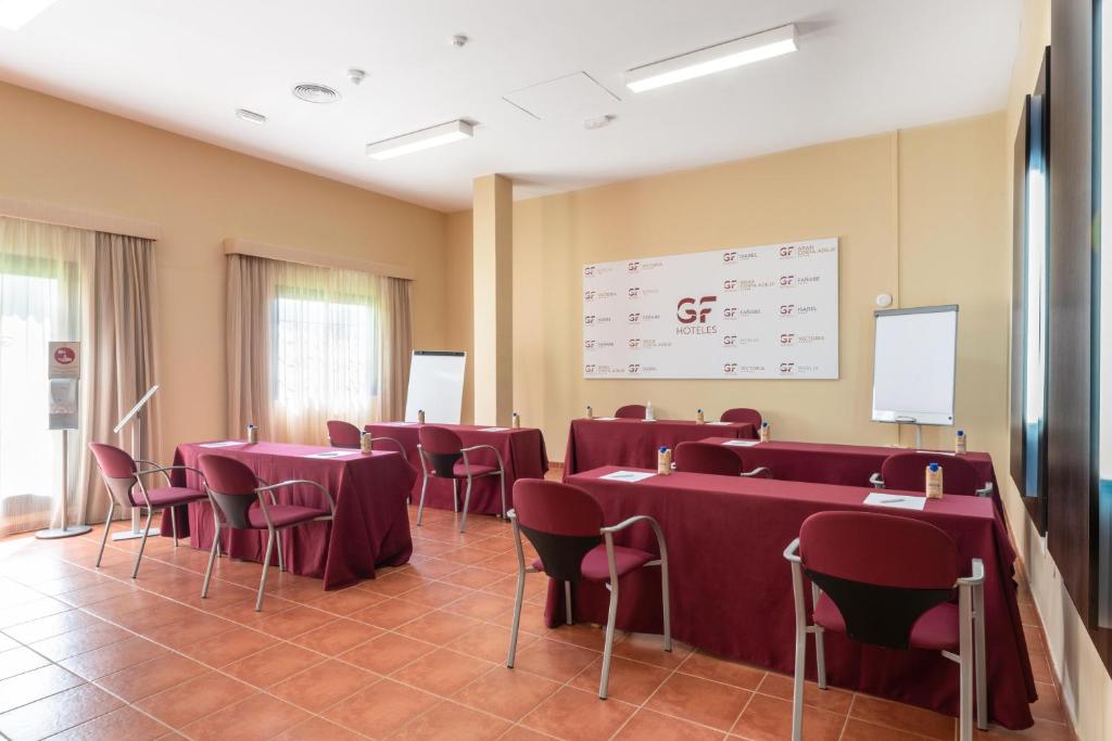 Konferenču telpa ar sarkaniem galdiem un krēsliem, kur ir pieejami rakstāmi un prezentācijas rīki. Telpa ir gaiša un plaša ar logiem, kas nodrošina dabisko apgaismojumu.