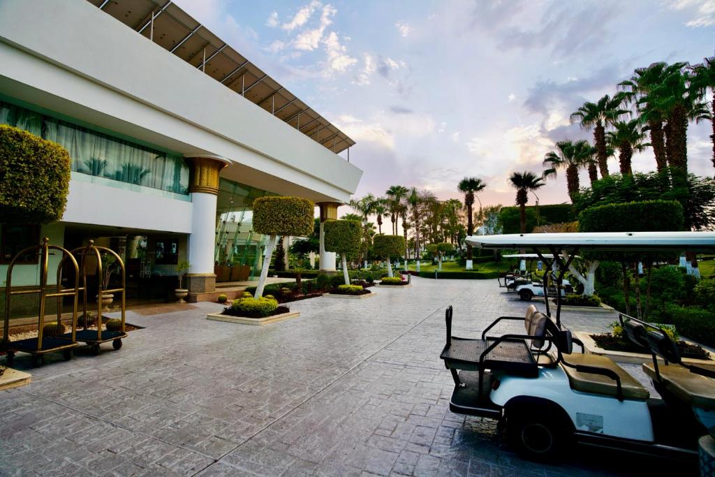 Mūsdienīgs viesnīcas ieeja ar golfa ratiņiem un greznām ainavām, kur redzami palmām un sakoptām krūmām. Gaisma un debesis rada relaksējošu atmosfēru.
