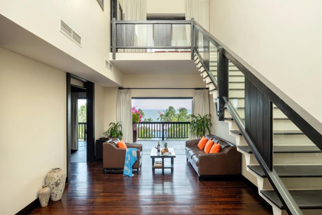 Mūsdienīgs viesistabas interjers ar diviem stāviem, stikla margām un lielu logu, kas piedāvā skatu uz dārzu. Telpā ir dīvāns ar spilveniem un eleganti dekori.