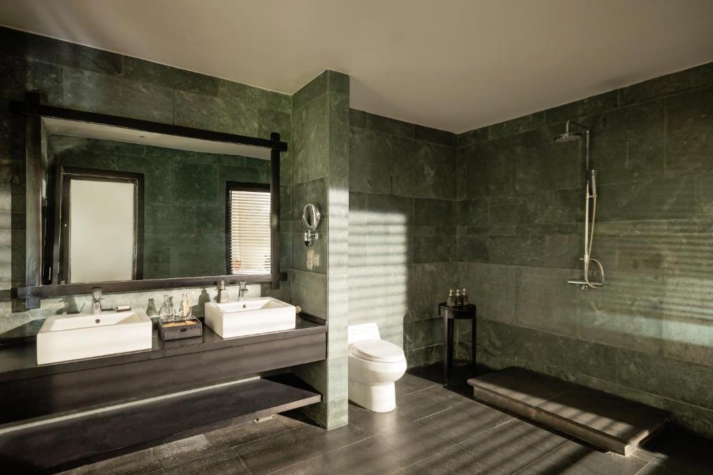 Mūsdienīgs vannas istabas interjers ar zaļām akmens sienām, divām izlietnēm un dušas kabīni. Gaisma ieplūst caur aizkariem, radot patīkamu atmosfēru.