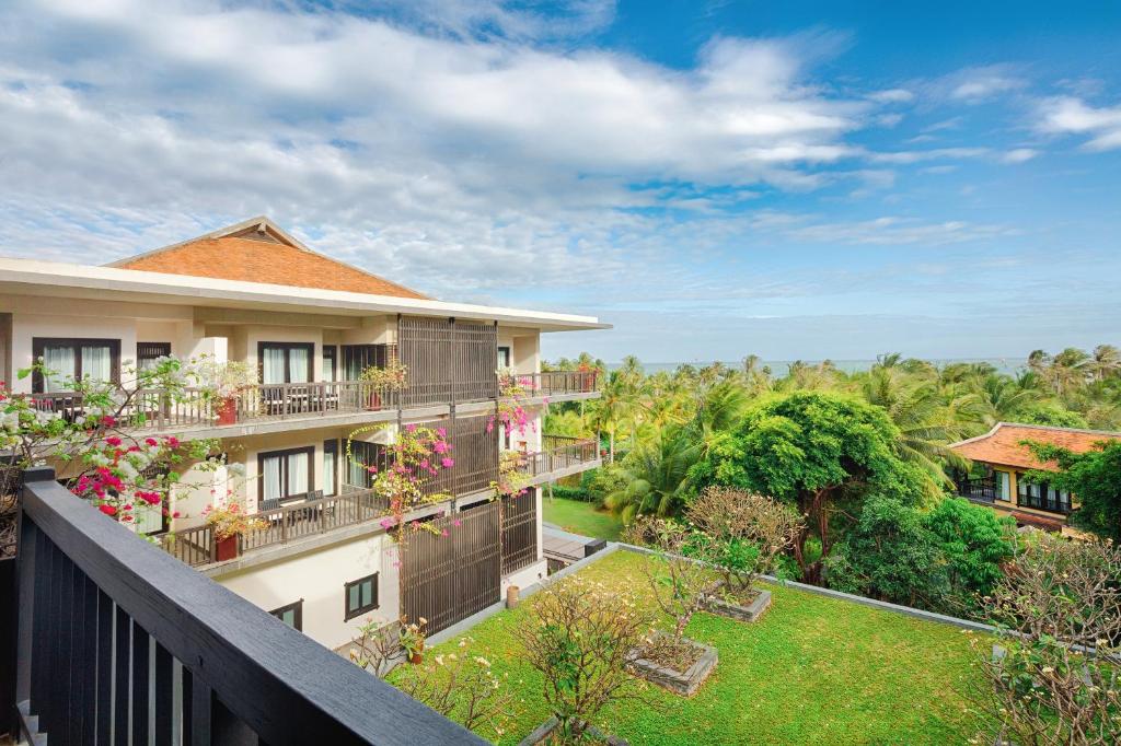 Viesnīcas skats ar zaļu dārzu un ziediem, kas rotā balkonus, pie jūras. Mierīga un tropiska atmosfēra ar zilo debesu fonu.
