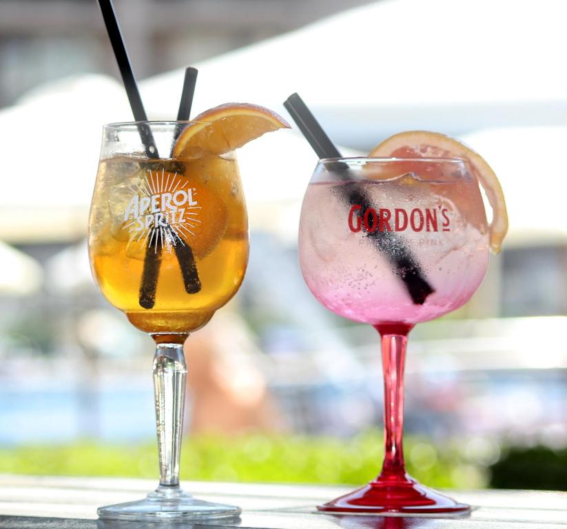 Divi dzērieni – oranžs Aperol Spritz un rozā Gordon's toniks – pasniegti elegantās glāzēs ar salmiņiem un garnējumu. Ideāls attēls vasaras atpūtai un dzērienu baudīšanai.