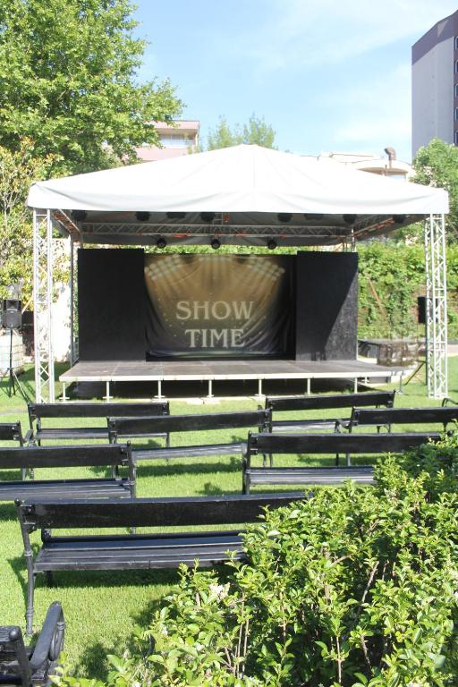 Āra skatuve ar telti, kas aprīkota ar gaismas tehniku un ar uzrakstu "SHOW TIME", priekšā rindas melnu solu. Apkārt zāle ar zaļām krūmiem.