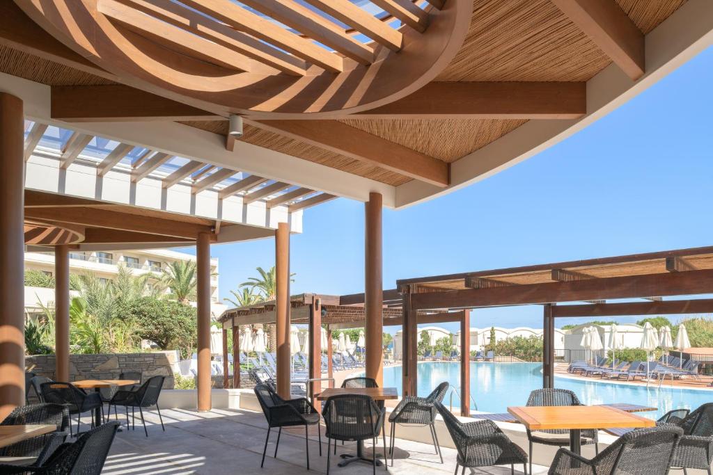 Atvērtā terase ar galdiem un krēsliem, kas izskatās uz peldbaseinu, apkārt ir tropu augi un saule spīd. Terase ir veidota ar koka konstrukcijām un caurspīdīgām jumta daļām, radot relaksējošu atmosfēru.