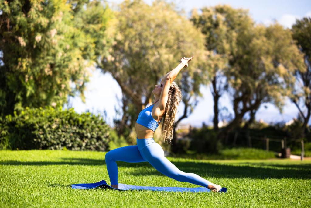 Sieviete veic jogas pozīciju zaļā zālienā, tērpta zilos sporta apģērbos, ar izstieptām rokām un relaksētu pozāciju. Apkārt redzama daba ar kokiem un zālienu.