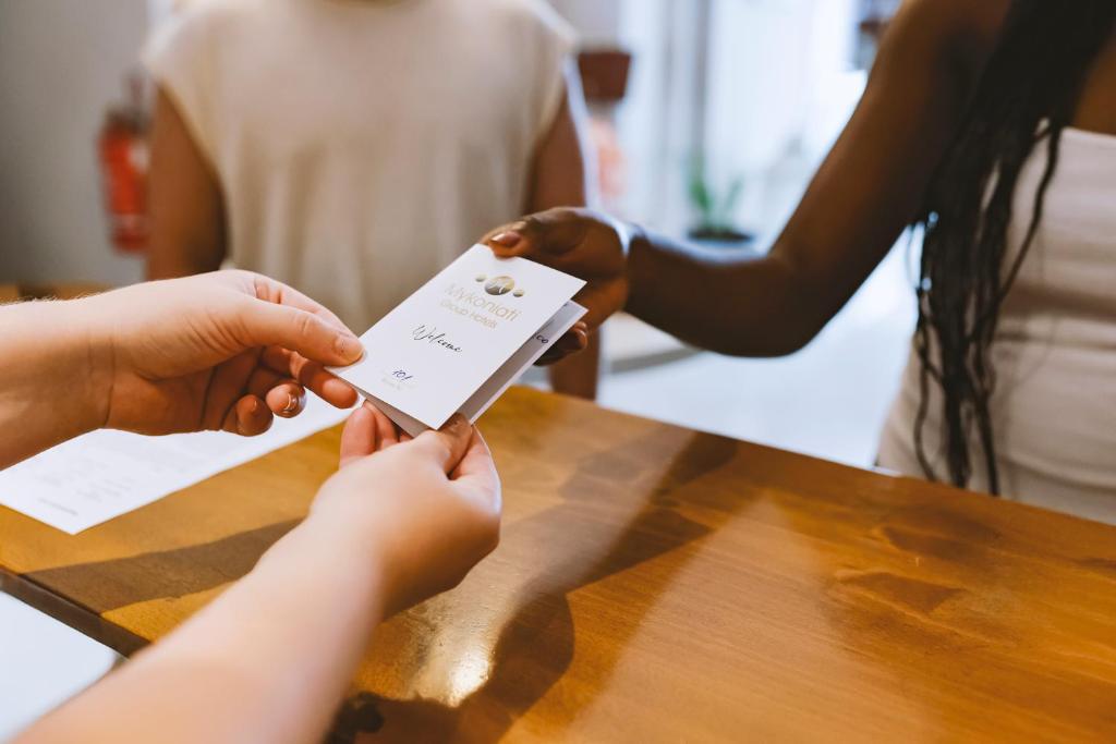 Rokas, kas nodod vizīti kādam citam, uz koka galda. Redzama arī daļa no citu cilvēku rokas un apkārtējās vides detaļas.