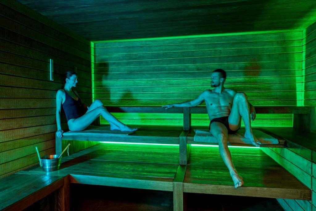 Divi cilvēki atpūšas mūsdienīgā saunā ar zaļganu apgaismojumu. Tiek attēloti relaksācijas brīži un veselīga dzīvesveida baudīšana.