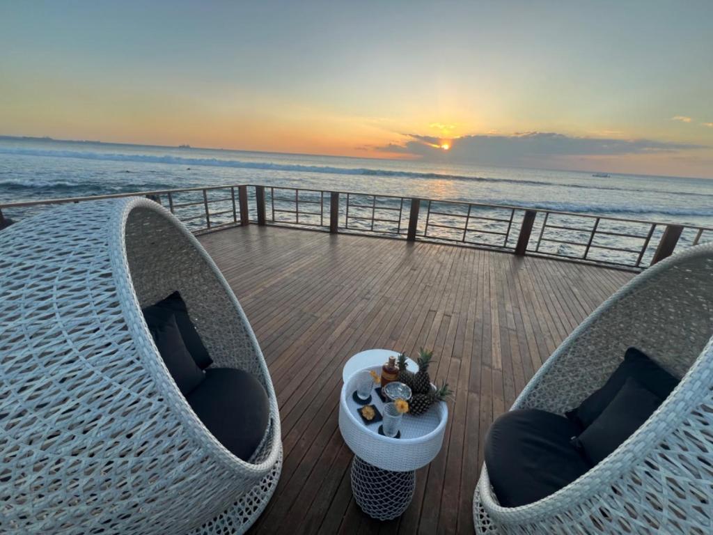 Romantiska atpūtas zona ar divām baltiem krēsliem un galdiņu, uz kura atrodas augļi un dzērieni, pie jūras ar saulrietu fona. Ideāla vieta relaksācijai un baudīšanai.