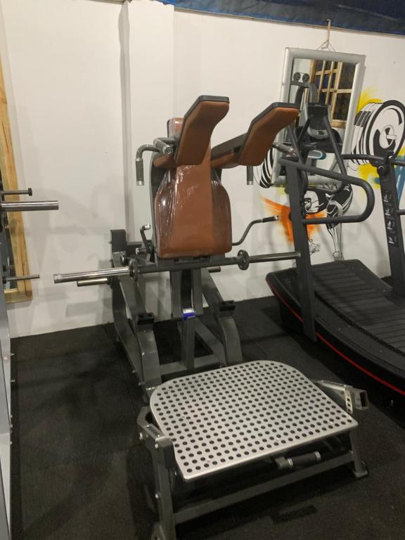 Vingrošanas aprīkojums ar brūnu sēdekli un regulējamu atzveltni, blakus esošs trenažieris un gumijas grīdas segums. Ideāli piemērots sporta zāles videi.