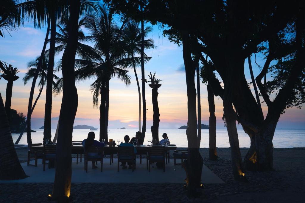 Saulrieta ainava pludmalē, kur cilvēki sēž pie galda zem palmām, baudot mierīgu mirkli. Fons piepildīts ar krāsainām debesīm un jūras ainavām.