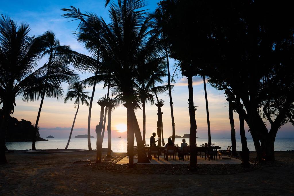Saule riet krastā, iepludinot silto gaismu starp palmām un kokiem, kur cilvēki bauda vakariņas pie galda. Klusa pludmale ar mierīgu jūru un tāliem kalniem.