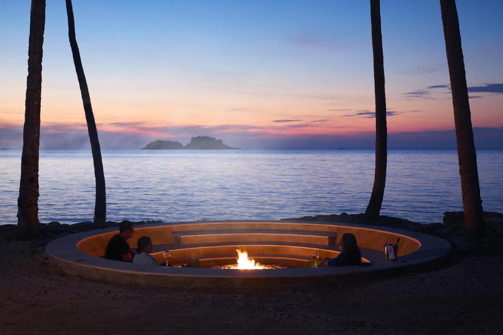 Romantisks skats uz saulrietu pie jūras ar ugunskuru un diviem cilvēkiem, kas izbauda mierīgu vakaru zem palmām.