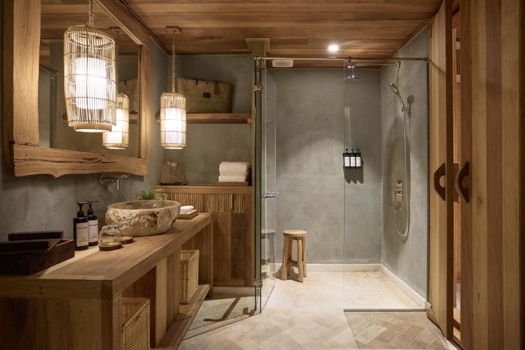Mūsdienīgs vannas istabas interjers ar koka elementiem, akmens izlietni un stikla dušu, radot siltu un relaksējošu atmosfēru. Apgaismojums ar rotājumiem piešķir telpai mājīgumu.