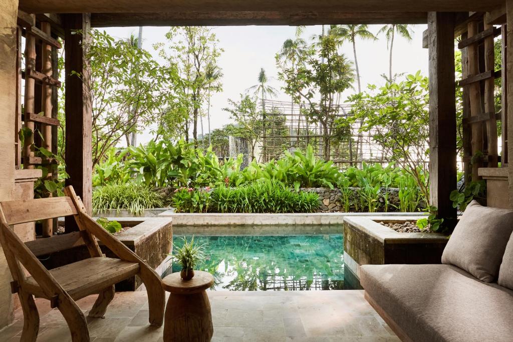 Mierīga ainava ar baseinu un tropisko veģetāciju, kas redzama no stilīgas atpūtas zonas. Koka mēbeles un zaļumi rada relaksējošu atmosfēru.