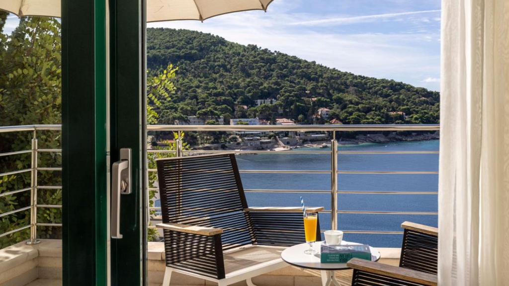 Balkons ar skatu uz jūru, kur divas krēsli un galds ar dzērienu, apkārt zaļi kalni un mājas.