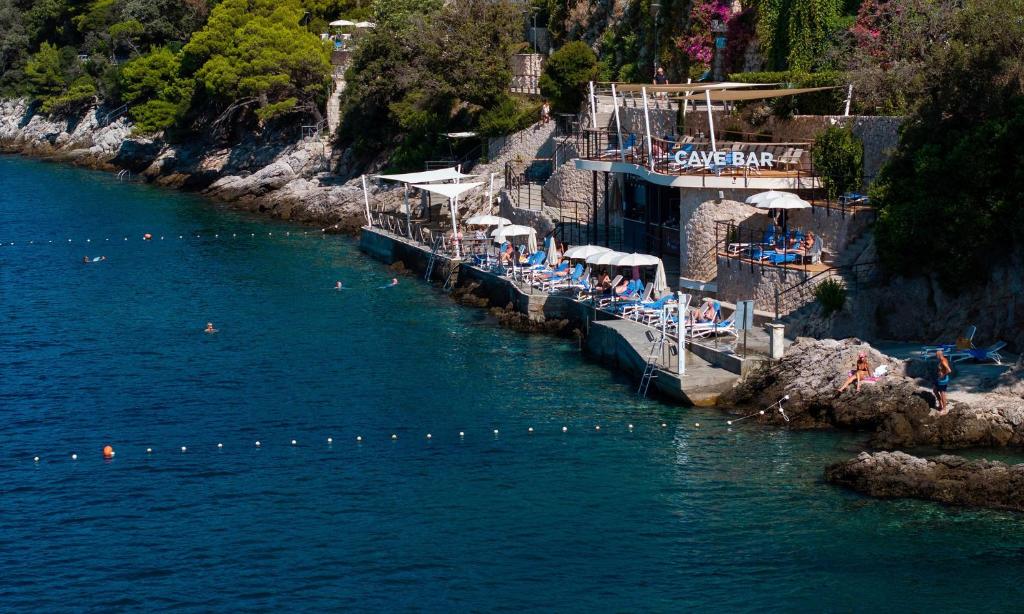 Kleita pludmales bārs pie jūras, kur apmeklētāji relaksējas uz sauļošanās krēsliem, baudot sauli un ūdeni. Fons ir klints un zaļi augi, kas rada mierīgu atmosfēru.