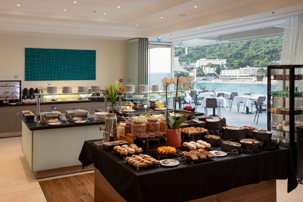 Bufete ar dažādiem ēdieniem un desertiem, kas izkārtoti uz galdiem, ar skatu uz jūru un zaļiem pakalniem.