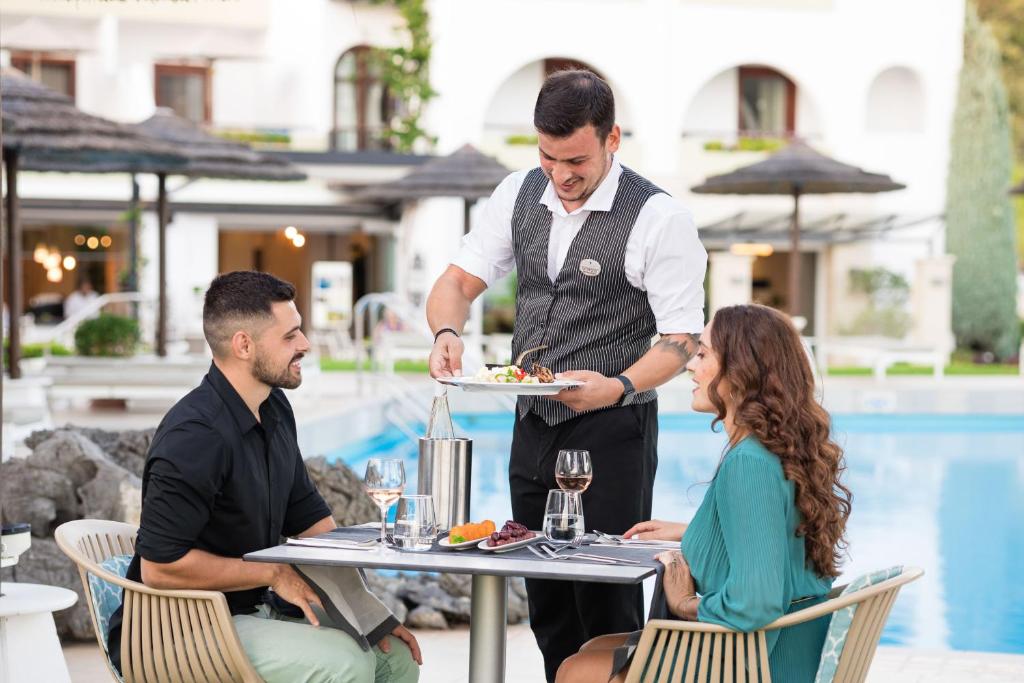 Romantiska vakariņas pie baseina, kur viesmīlis pasniedz ēdienu pāri, kam ir patīkama saruna. Elegantā atmosfērā redzami galda piederumi un dzērieni.