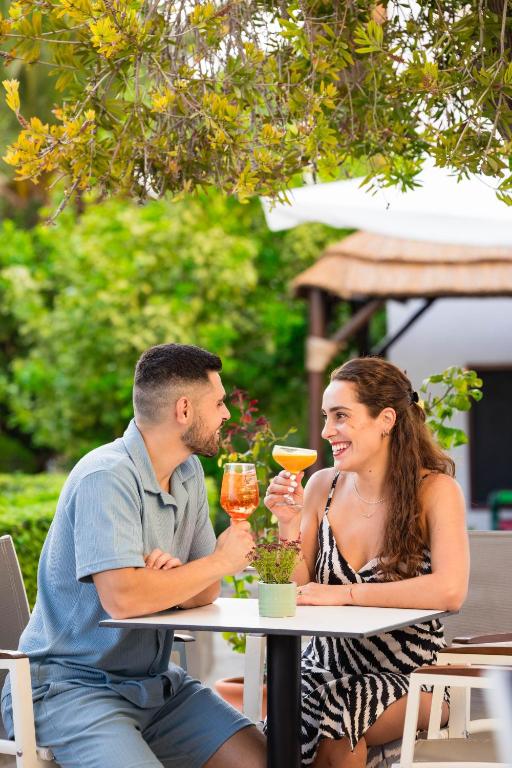 Pāris, sēžot pie galda, baudā atspirdzinošus kokteiļus, smaidot un skatoties viens uz otru, apkārt plaukst zaļa dārza vide.