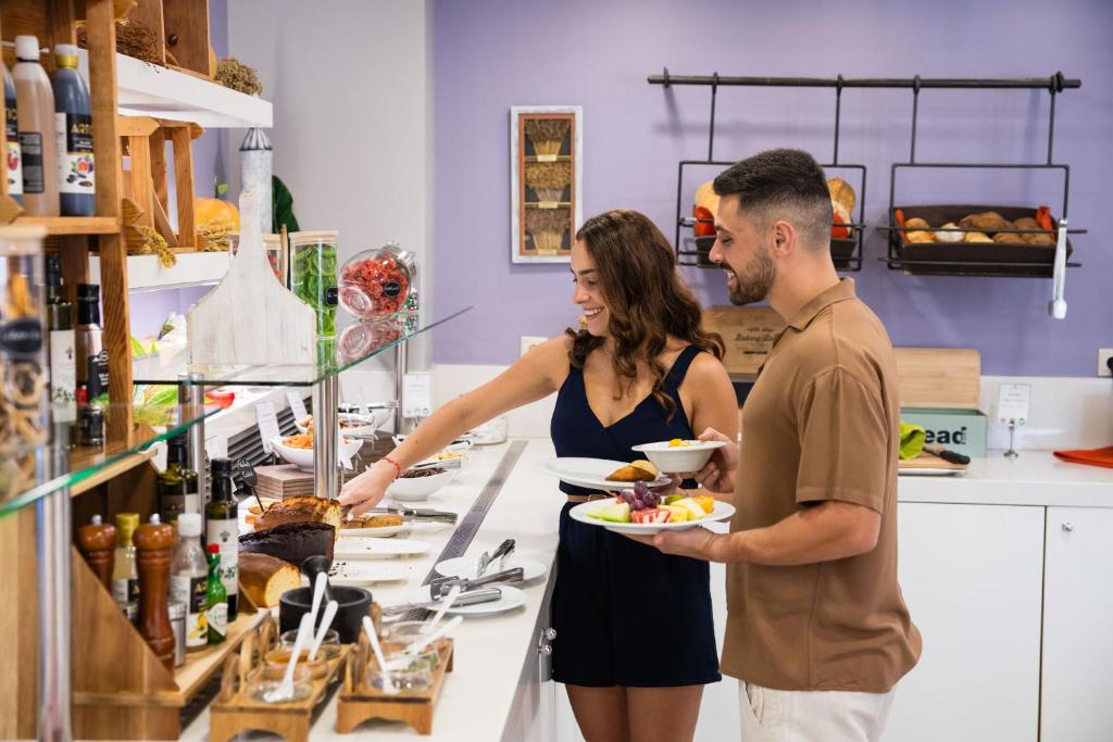 Divi cilvēki, vīrietis un sieviete, stāv pie pašapkalpošanās bufetes, izvēloties pārtiku no dažādiem ēdieniem un dzērieniem, kamēr fons ir rotāts ar maizes groziem un dekoratīvām detaļām.