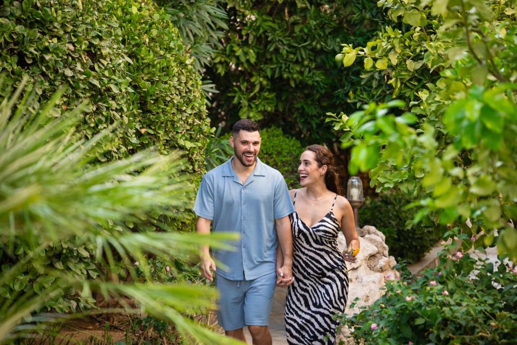 Pāris, turot rokas, staigā pa zaļu dārzu, apņemti ar krūmiem un augiem, izskatās laimīgi un smaidīgi.