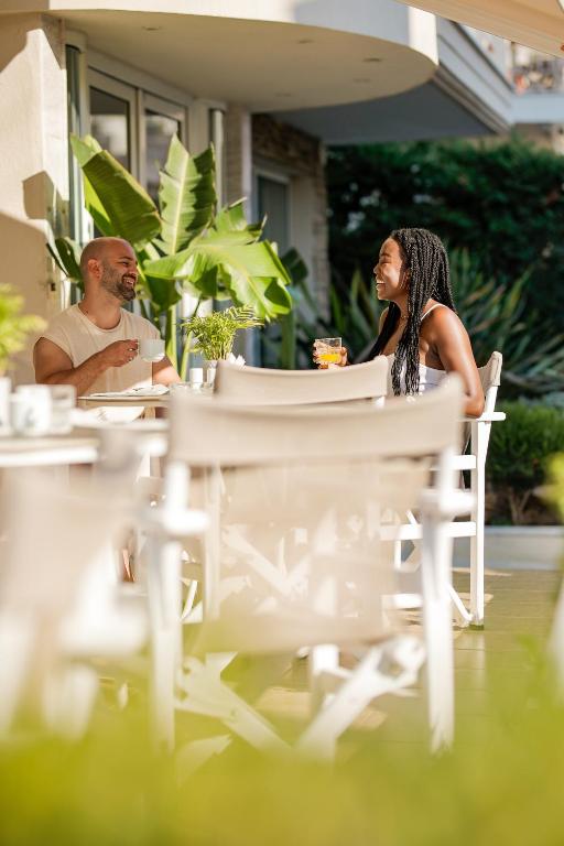 Divi cilvēki, baudot dzērienus un sarunājoties, atpūšas terasē, apstādījumos ar zaļām augiem. Gaisīgs un saulains brīdis, kas rada draudzīgu atmosfēru.