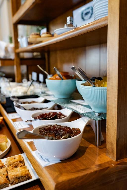 Bagātīgs brokastu bufetes galdiņš ar dažādām mērēm un uzkodām, kas pasniegtas baltās traukos. Apakšā redzami arī saldumi.