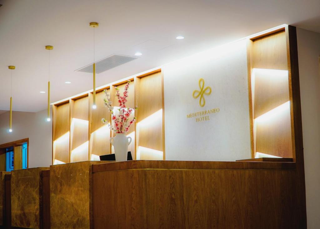 Mūsdienīga viesnīcas reģistratūra ar siltu apgaismojumu un dekoratīvu ziedu kompozīciju. Viesnīcas nosaukums "Mediterraneo Hotel" ir redzams uz sienas.