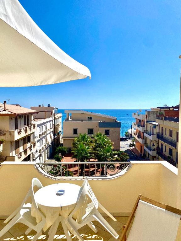 Balcony with a view of the sea, featuring a table and chairs, surrounded by buildings and palm trees under a clear blue sky.