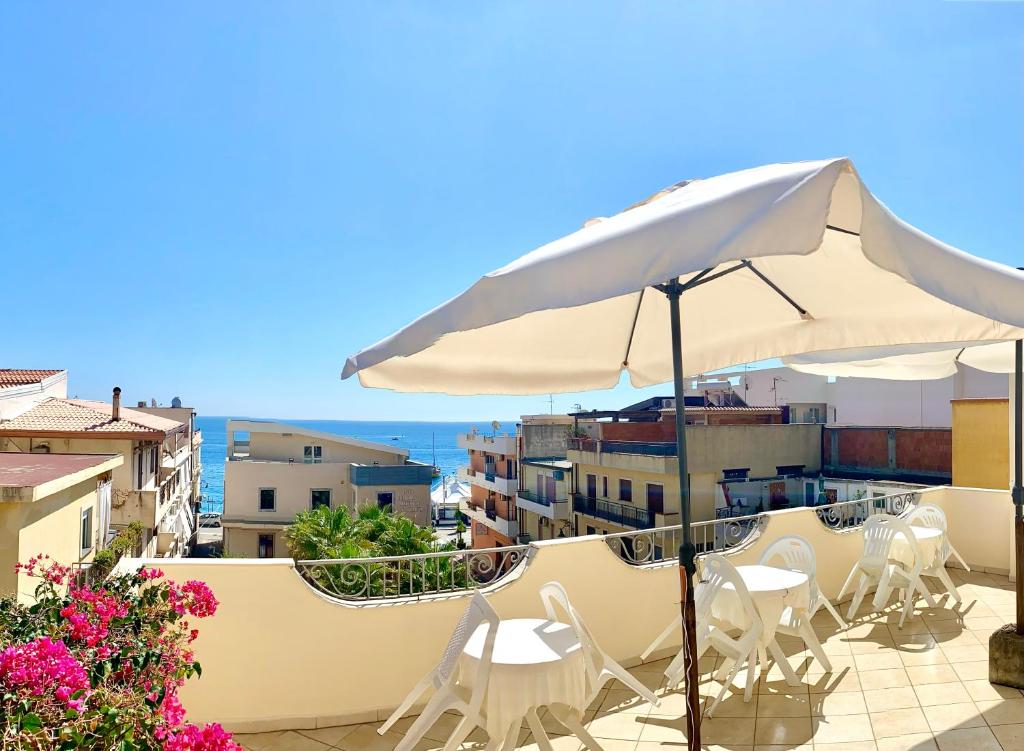 Skats no terases uz jūru ar baltiem krēsliem un saulessargiem, apkārtējās ēkas un rozā ziedi.