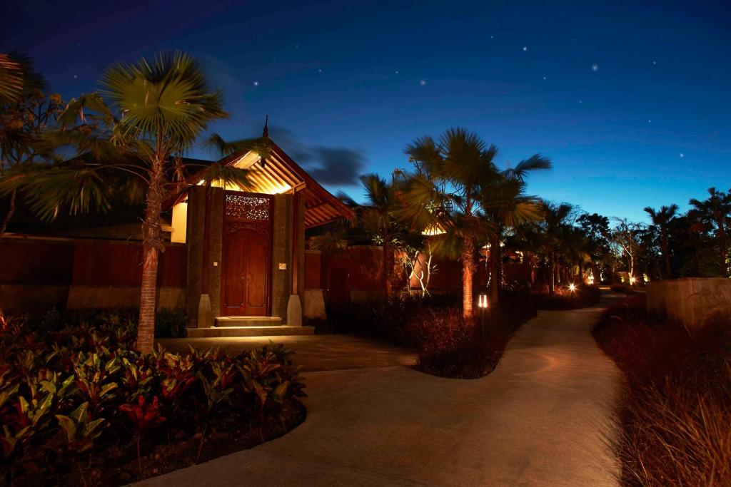 Tumsains apgaismotais ceļš ved uz elegantu māju ar izsmalcinātu durvju dizainu, ko ieskauj tropiskas palmas un ziedi, radot mierīgu un idillisku atmosfēru.